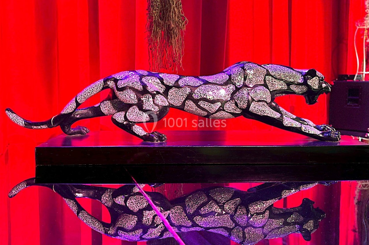 Statue de panthère incrustée de cristaux, posée sur un socle noir, devant un rideau rouge avec un reflet sur une surface…