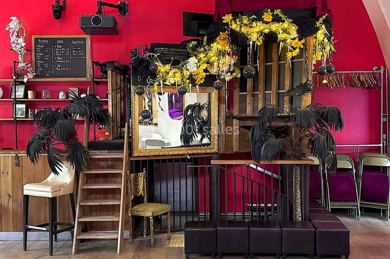 Décor intérieur avec estrade en bois, miroirs, fleurs jaunes et blanches, et plantes noires dans une salle rouge.