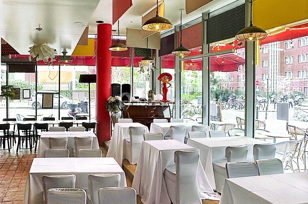 Salle de restaurant lumineuse avec des tables recouvertes de nappes blanches, grandes baies vitrées et décor coloré.
