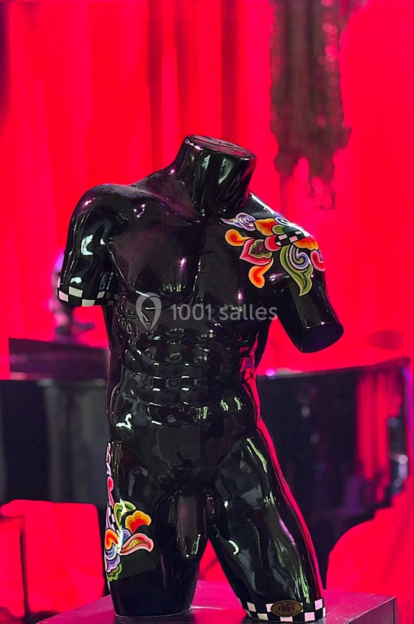 Buste masculin noir sans tête, orné de motifs colorés, exposé devant un rideau rouge et un piano en arrière-plan.