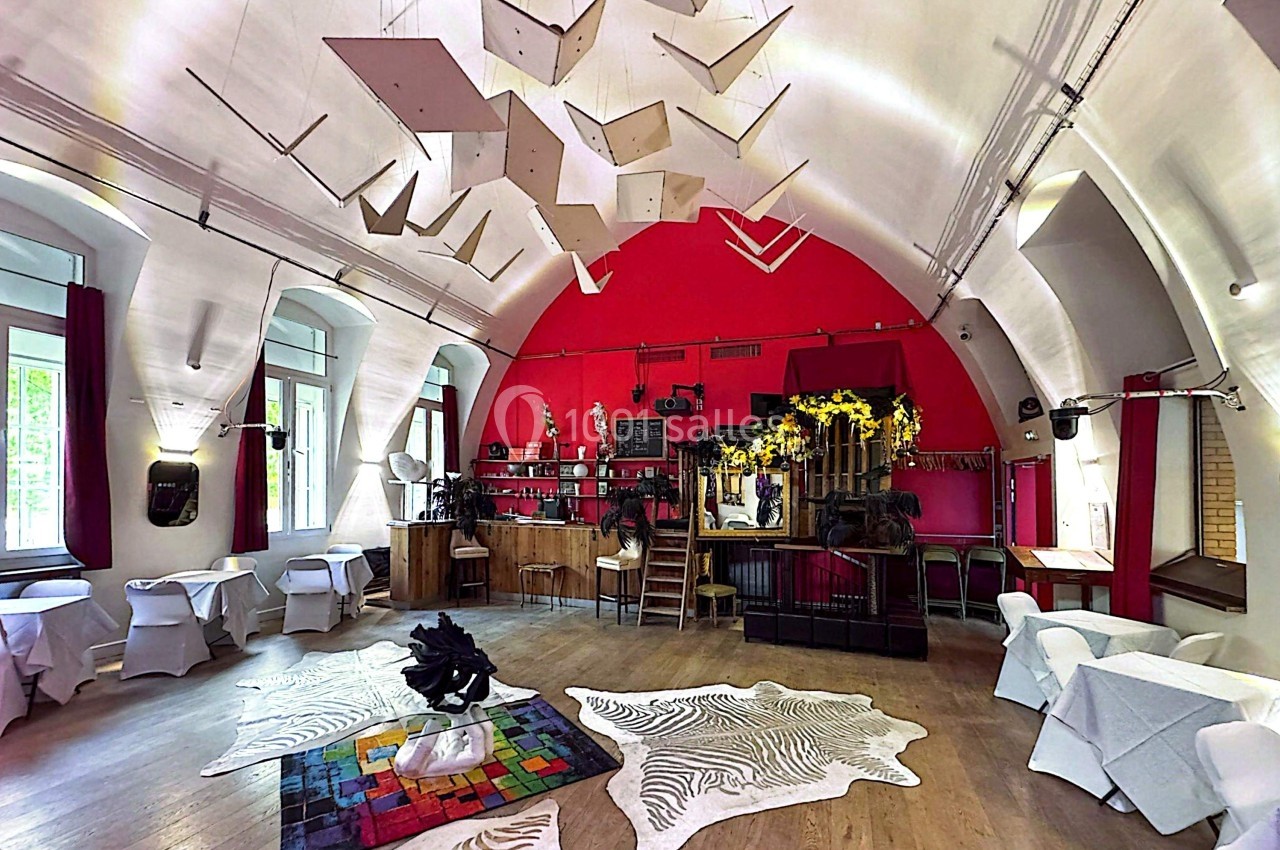 Salle lumineuse avec plafond voûté, décorations suspendues, tables dressées et espace bar en fond.