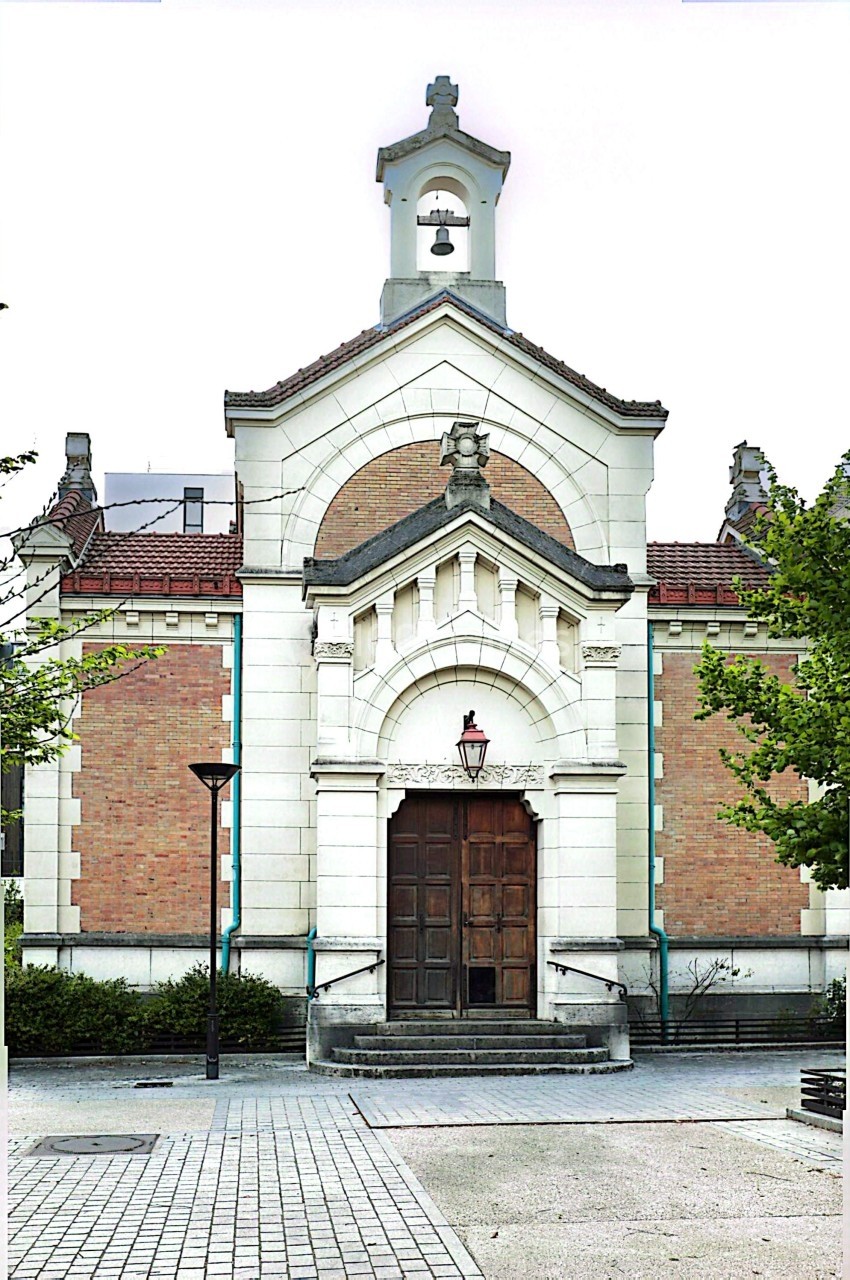 Façade d'un bâtiment en briques et pierre avec une porte en bois, un clocher et des arbres au premier plan.