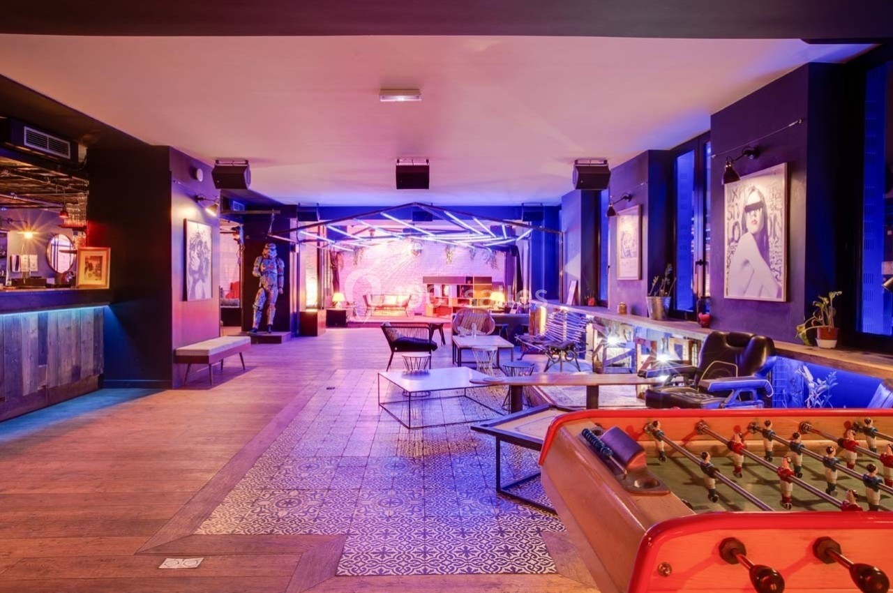 Salle moderne avec éclairage coloré, mobilier varié, baby-foot au premier plan et décoration artistique aux murs.