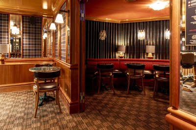 Salle avec tables et chaises en bois, banquettes rouges, murs à motif écossais et éclairage tamisé.