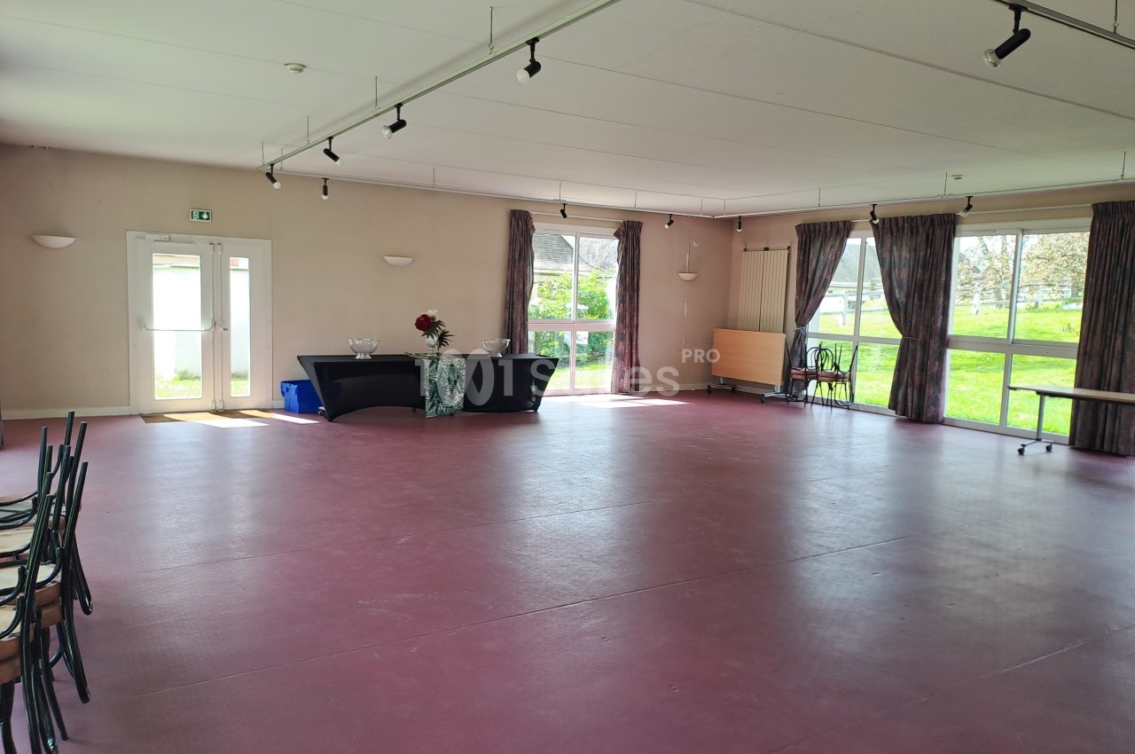 Salle vide avec sol rouge, grandes fenêtres laissant entrer la lumière, tables et chaises empilées sur le côté.