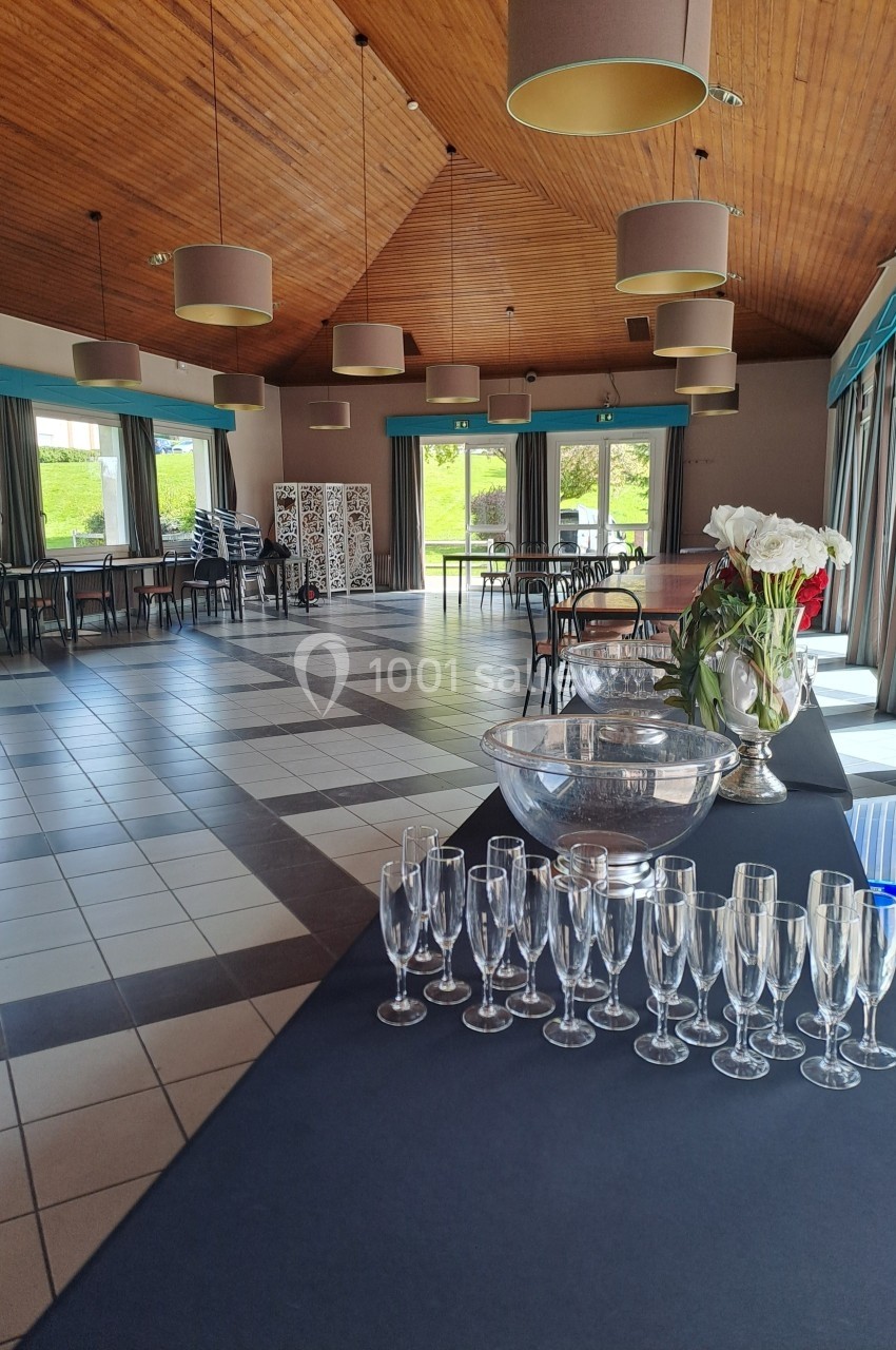 Salle lumineuse avec plafond en bois, tables espacées et rangées, flûtes à champagne alignées sur une table au premier plan.