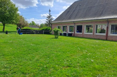 Pelouse verte entourée de bâtiments en briques et crépi, avec arbres et allée en béton sous un ciel dégagé.