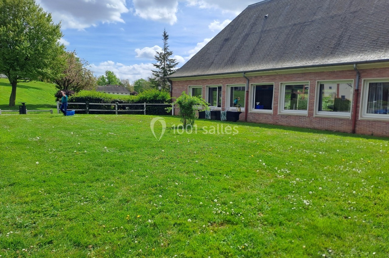 Pelouse verte avec quelques fleurs, bâtiment en briques avec fenêtres à droite, arbres et ciel partiellement nuageux en…