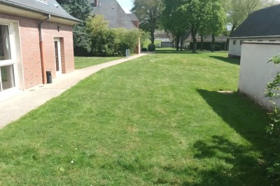 Pelouse verte entourée de bâtiments en briques et crépi, avec arbres et allée en béton sous un ciel dégagé.