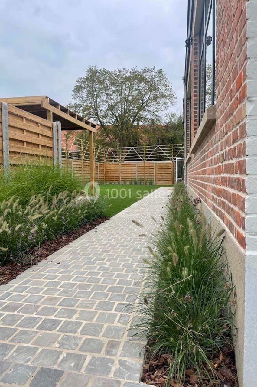 Allée en pavés bordée de plantes, menant à un jardin clôturé avec pergola en bois et pelouse.