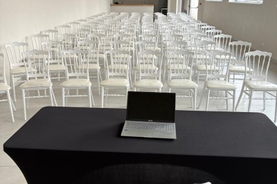 Salle lumineuse avec des rangées de chaises blanches alignées, plafond avec poutres apparentes et décor floral suspendu.