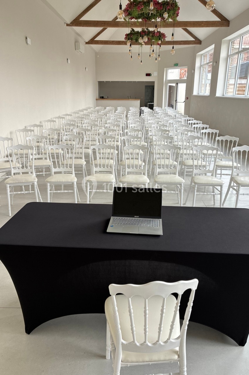 Salle de conférence lumineuse avec des rangées de chaises blanches, une table noire et un ordinateur portable.