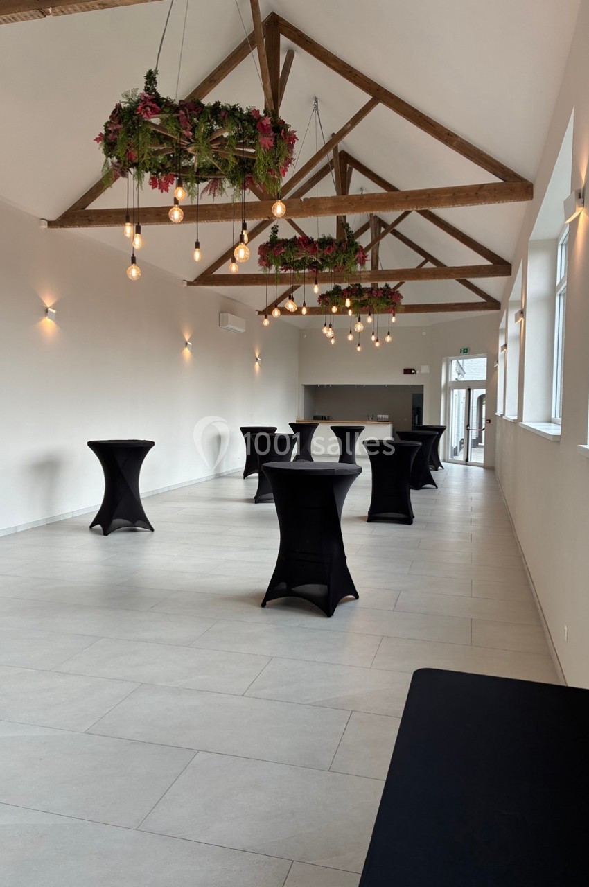 Salle lumineuse avec poutres apparentes, tables hautes noires et suspensions décorées de plantes et ampoules.