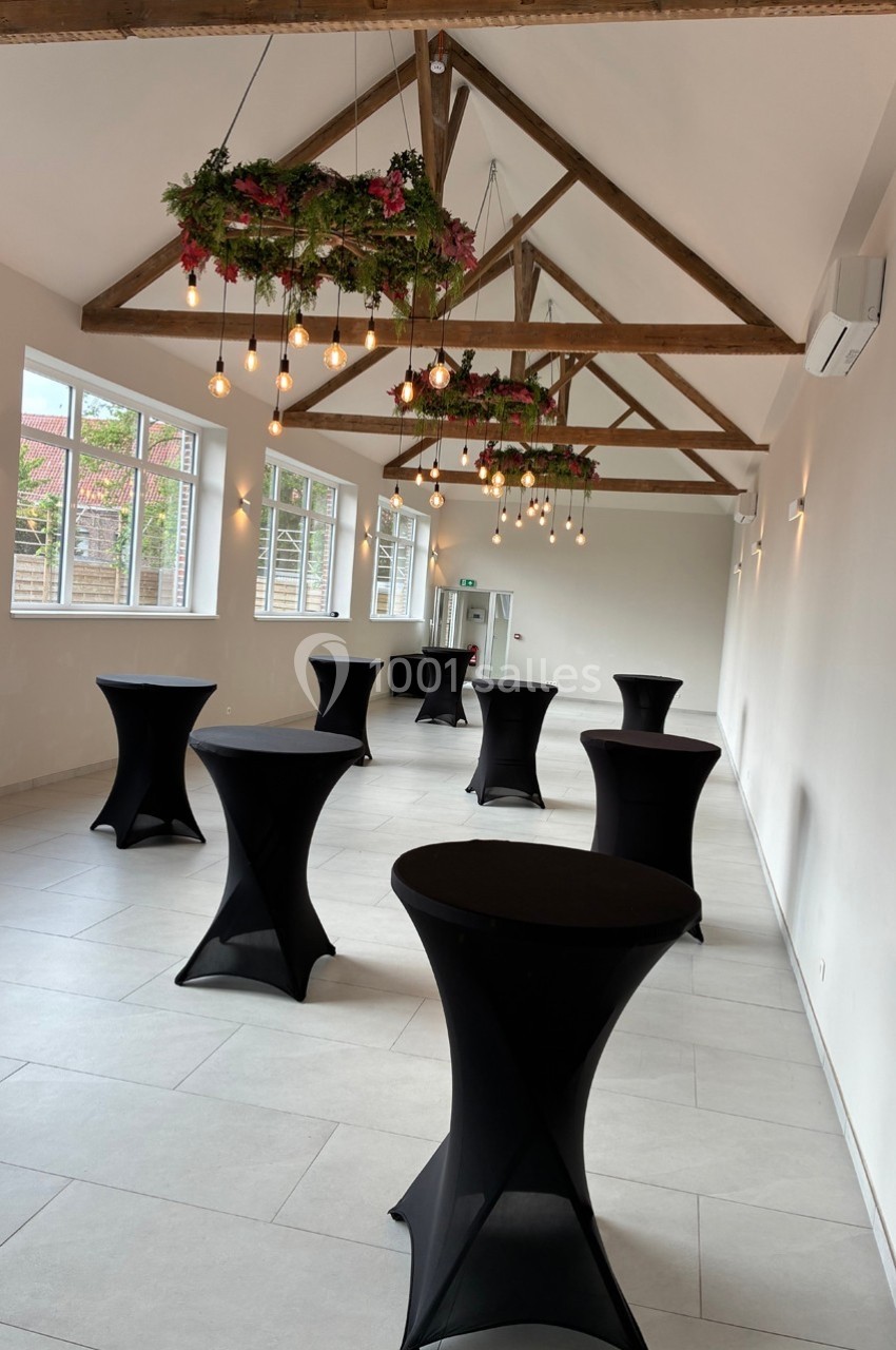 Salle lumineuse avec poutres apparentes, tables hautes noires et décorations suspendues en verdure et ampoules.