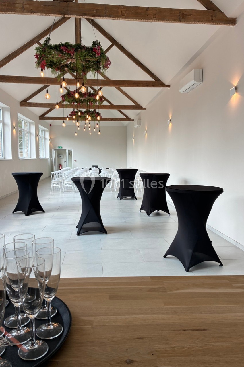 Salle lumineuse avec poutres apparentes, tables hautes noires, décor floral suspendu et verres à champagne au premier plan.