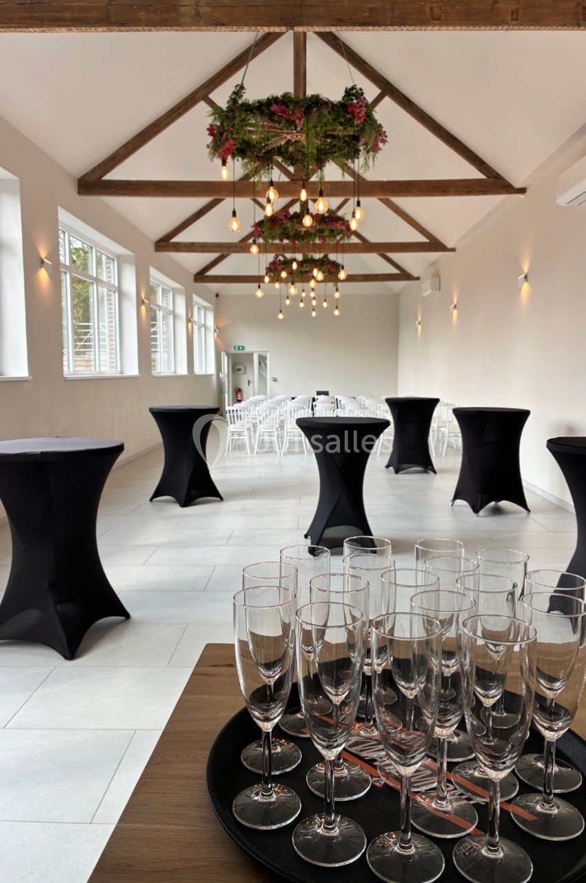 Salle lumineuse avec tables hautes noires, chaises blanches alignées et verres à champagne sur un plateau au premier plan.