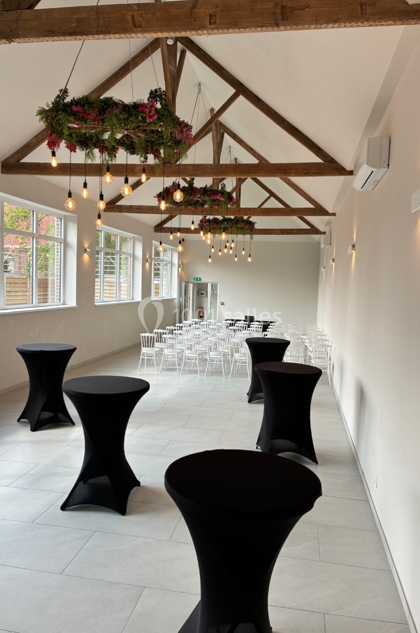 Salle lumineuse avec poutres apparentes, tables hautes noires au premier plan et chaises blanches disposées au fond.