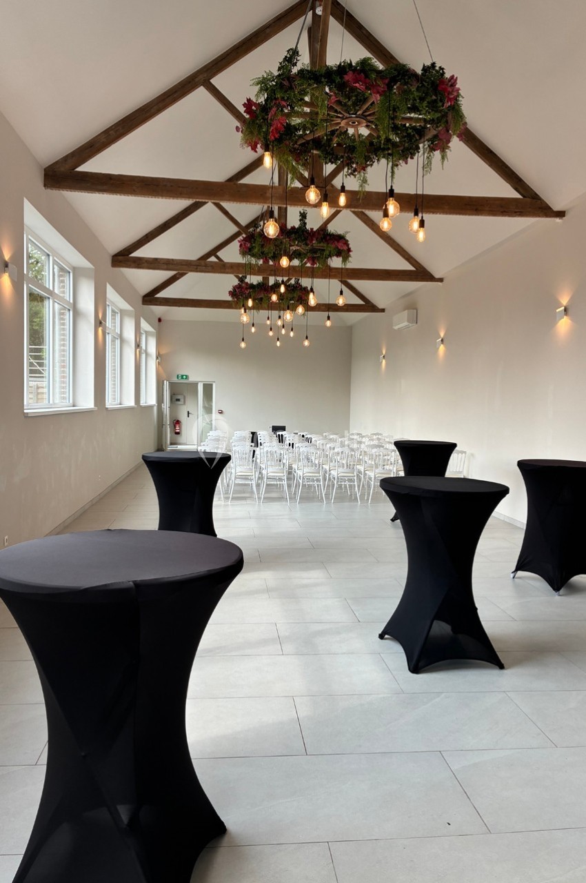 Salle lumineuse avec tables hautes noires, chaises blanches alignées au fond et suspensions décorées de feuillage.