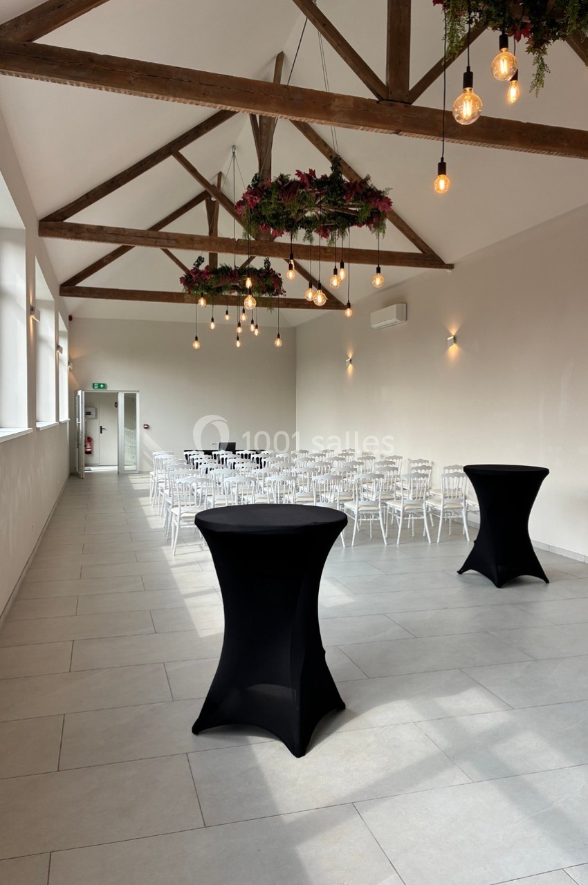 Salle lumineuse avec poutres apparentes, chaises blanches alignées, tables hautes noires et décorations suspendues.