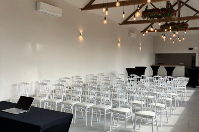 Salle lumineuse avec des rangées de chaises blanches alignées, plafond avec poutres apparentes et décor floral suspendu.