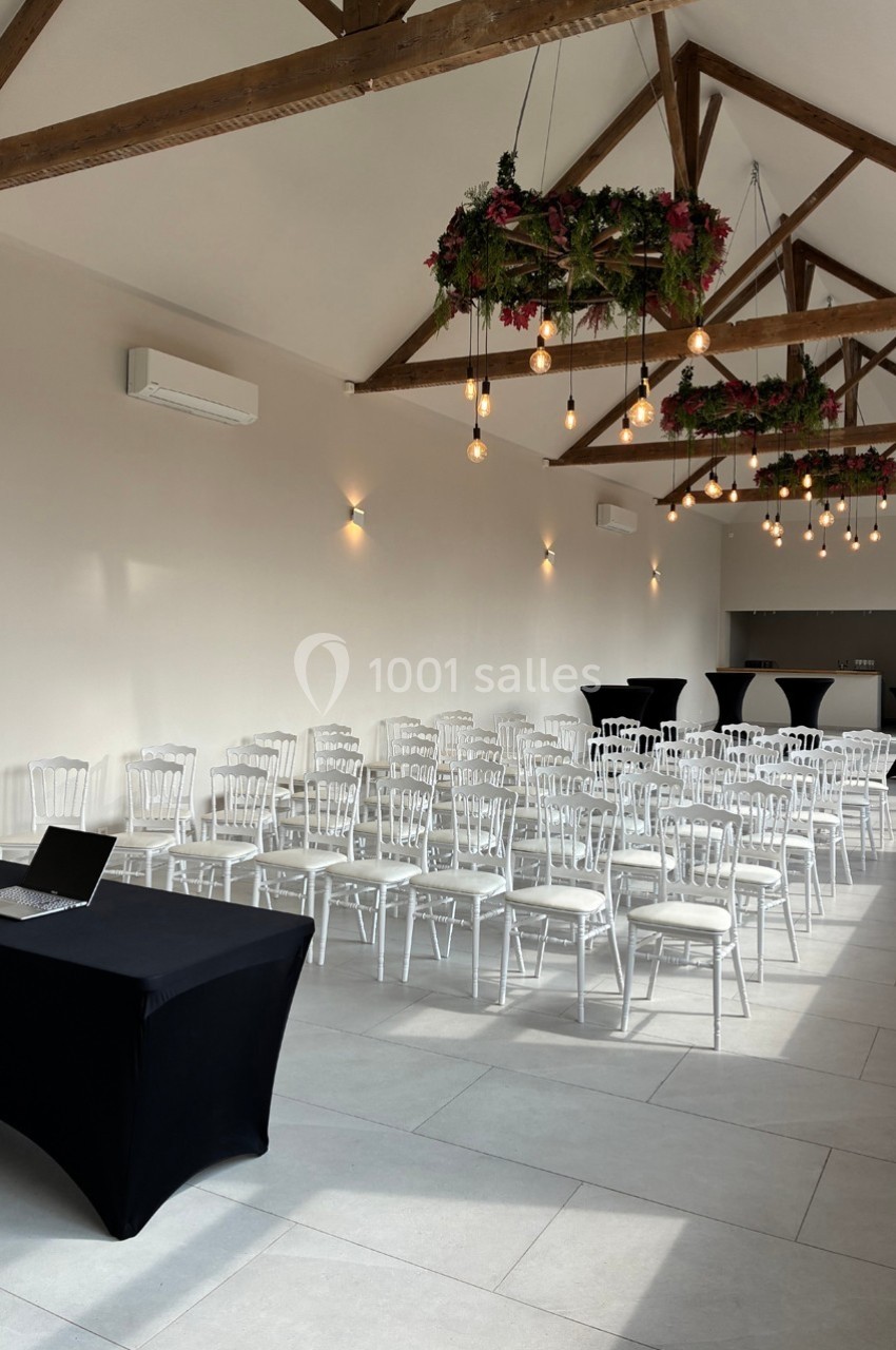 Salle lumineuse avec des chaises blanches alignées, des suspensions végétales et une table noire avec un ordinateur.