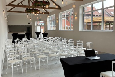 Salle lumineuse avec des rangées de chaises blanches alignées, plafond avec poutres apparentes et décor floral suspendu.