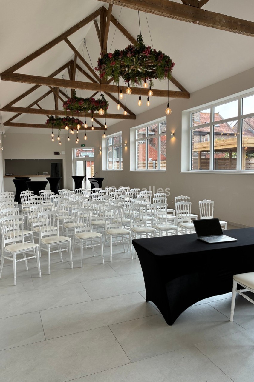 Salle lumineuse avec des chaises blanches alignées, une table noire avec un ordinateur et des décorations suspendues.