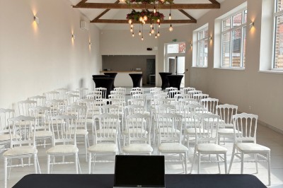 Salle lumineuse avec des rangées de chaises blanches alignées, plafond avec poutres apparentes et décor floral suspendu.
