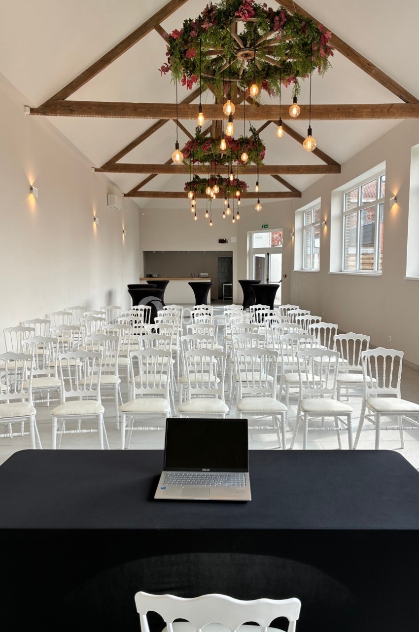 Salle lumineuse aménagée pour une conférence, avec chaises blanches alignées, table noire et ordinateur portable au premier…