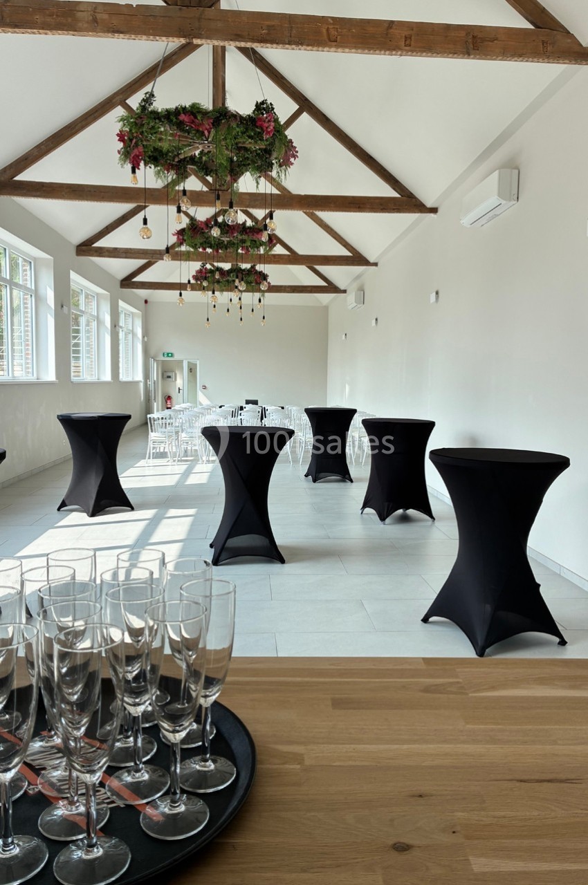 Salle lumineuse avec tables hautes noires, chaises blanches, verrière et décoration florale suspendue.