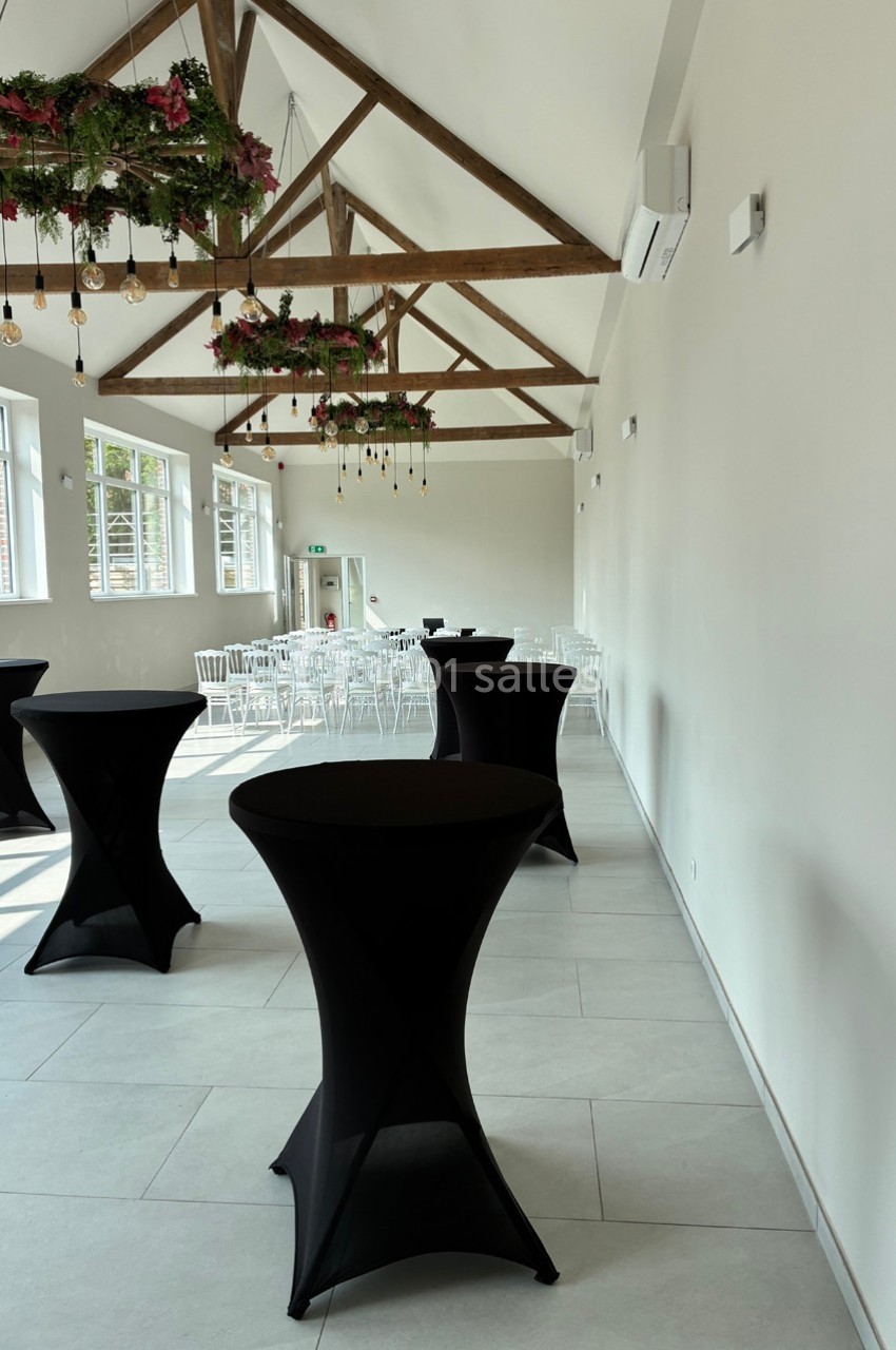 Salle lumineuse avec poutres apparentes, tables hautes noires et chaises blanches alignées près des fenêtres.