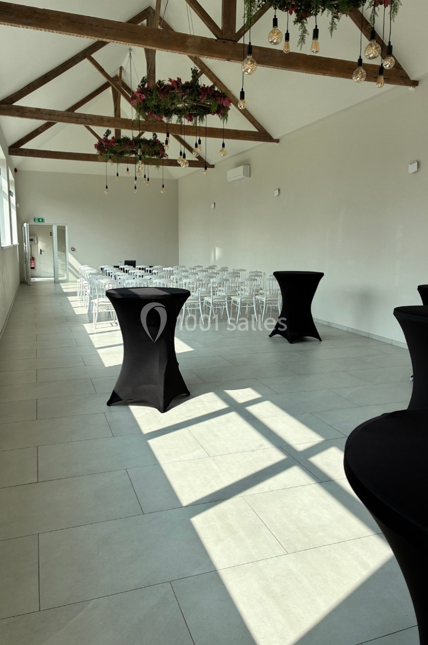 Salle lumineuse avec poutres apparentes, chaises blanches alignées, tables hautes noires et décor floral suspendu.