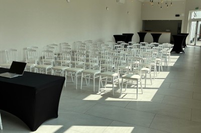 Salle lumineuse avec des rangées de chaises blanches alignées, plafond avec poutres apparentes et décor floral suspendu.
