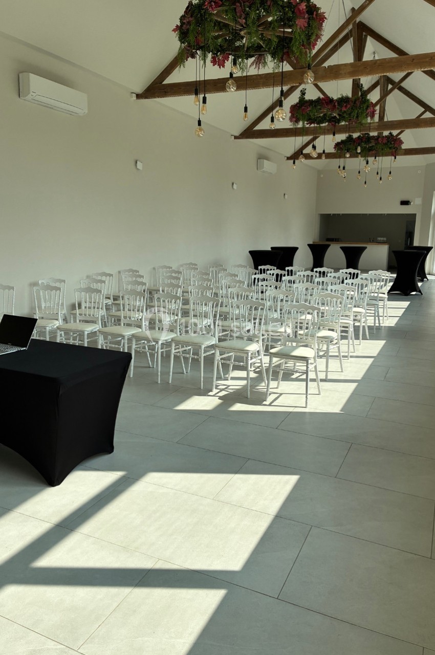 Salle lumineuse aménagée avec des chaises blanches alignées, une table noire et des décorations suspendues.