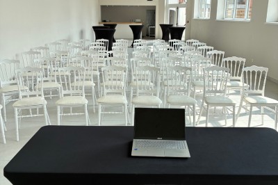 Salle lumineuse avec des rangées de chaises blanches alignées, plafond avec poutres apparentes et décor floral suspendu.