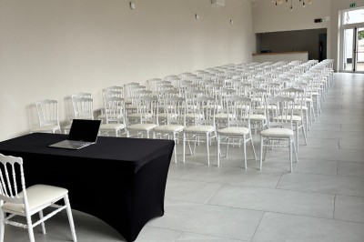 Salle lumineuse avec des rangées de chaises blanches alignées, plafond avec poutres apparentes et décor floral suspendu.