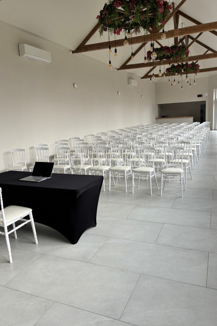 Salle lumineuse aménagée avec des rangées de chaises blanches, une table noire avec un ordinateur portable au premier plan.