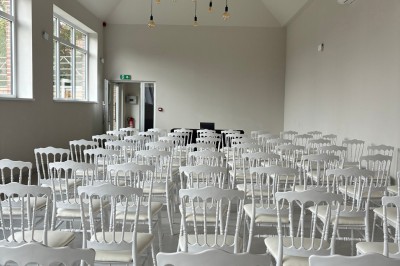 Salle lumineuse avec des rangées de chaises blanches alignées, plafond avec poutres apparentes et décor floral suspendu.