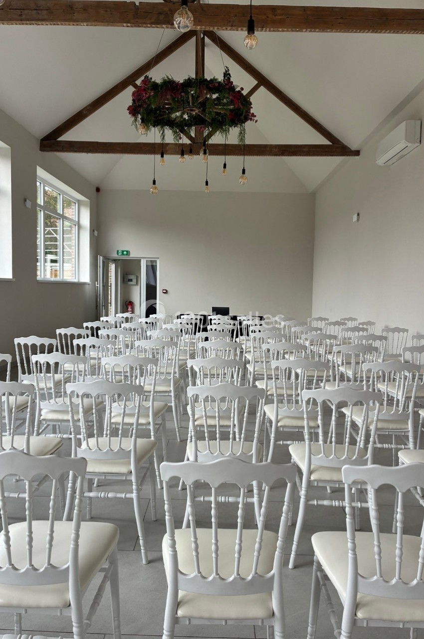 Salle lumineuse avec des chaises blanches alignées, un plafond en poutres apparentes et une décoration florale suspendue.