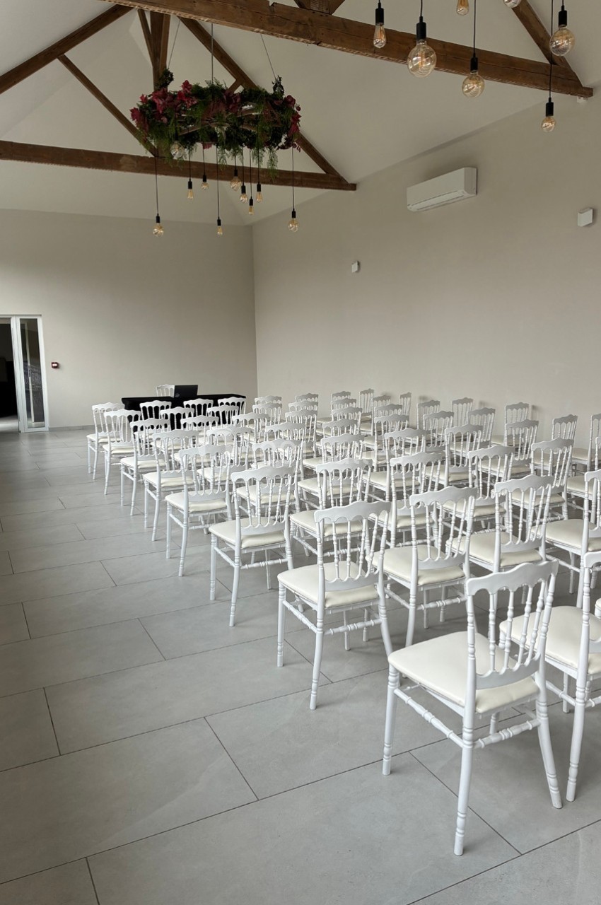 Salle lumineuse avec des chaises blanches alignées, plafond en poutres apparentes et décoration florale suspendue.