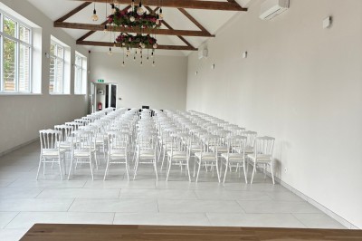 Salle lumineuse avec des rangées de chaises blanches alignées, plafond avec poutres apparentes et décor floral suspendu.