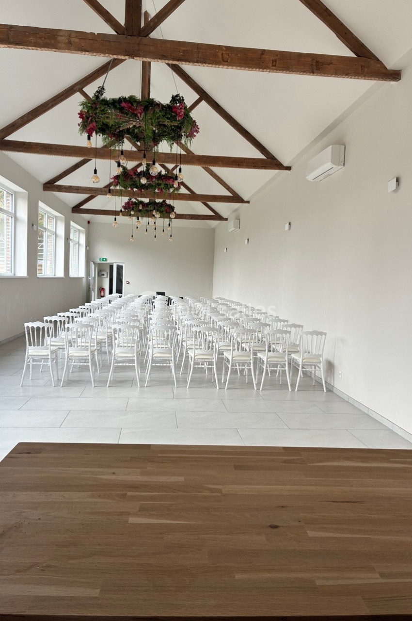 Salle lumineuse avec des chaises blanches alignées, plafond en poutres apparentes et décorations florales suspendues.