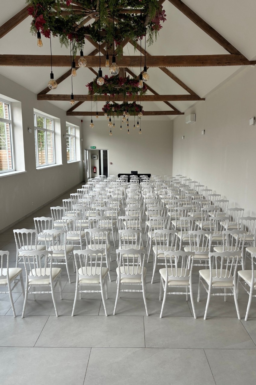 Salle lumineuse avec des rangées de chaises blanches alignées sous un plafond en poutres apparentes décoré de guirlandes.