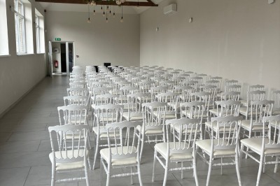 Salle lumineuse avec des rangées de chaises blanches alignées, plafond avec poutres apparentes et décor floral suspendu.