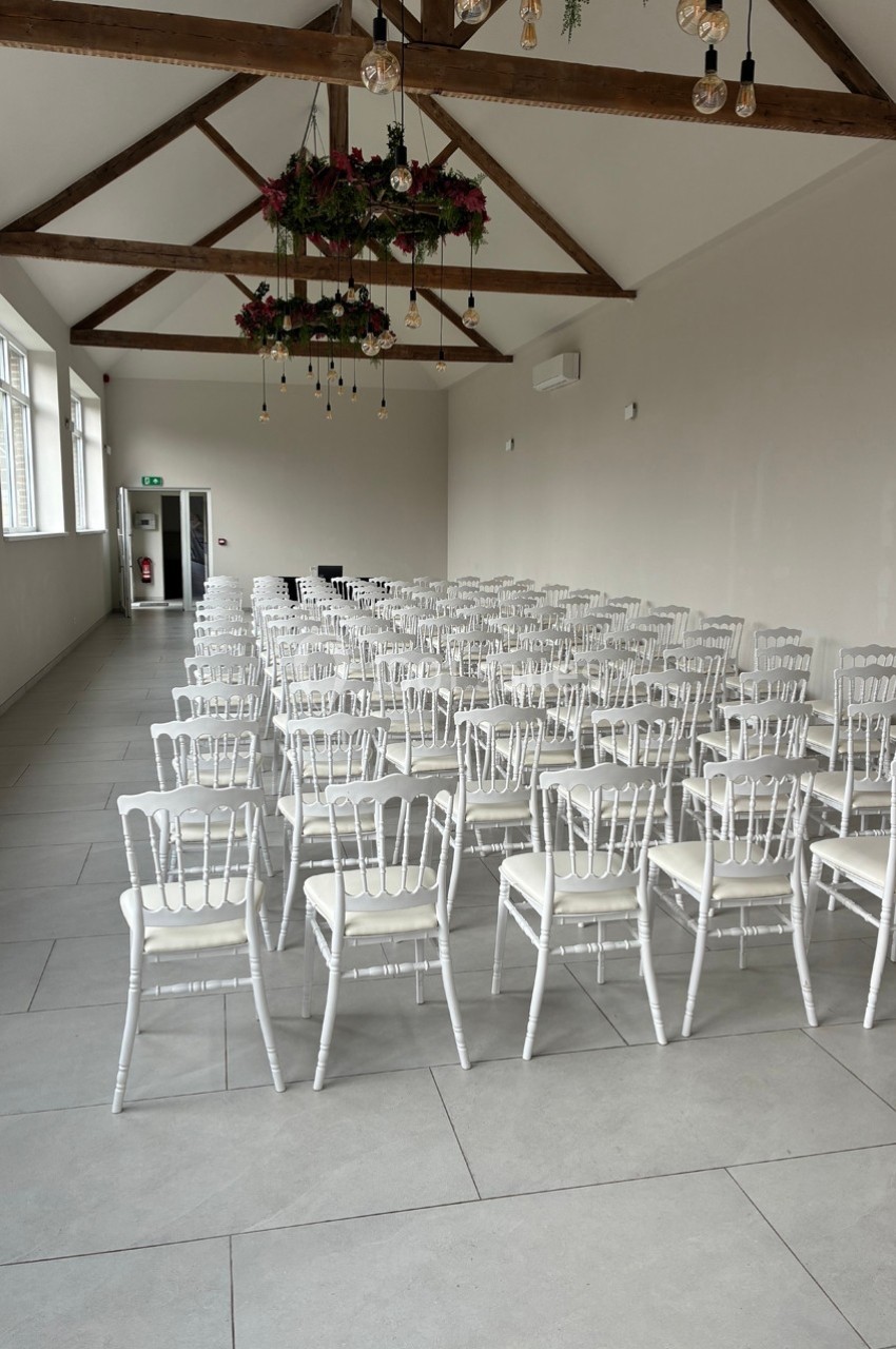 Salle lumineuse avec des rangées de chaises blanches alignées, plafond avec poutres apparentes et décor floral suspendu.