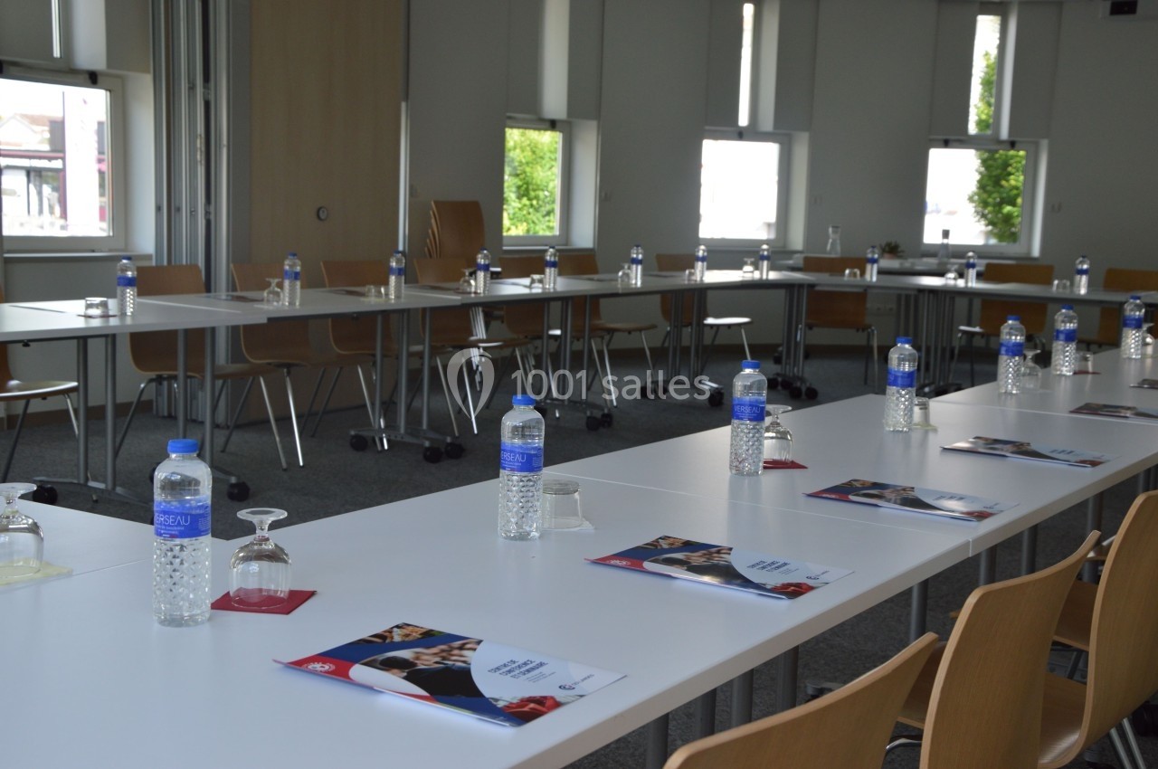 Salle de réunion lumineuse avec des tables en U, des bouteilles d'eau, des verres et des brochures disposés.