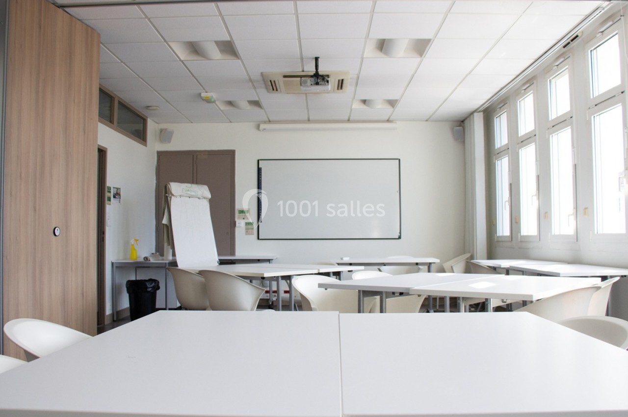 Salle de classe lumineuse avec tables blanches, chaises, tableau blanc et grandes fenêtres.