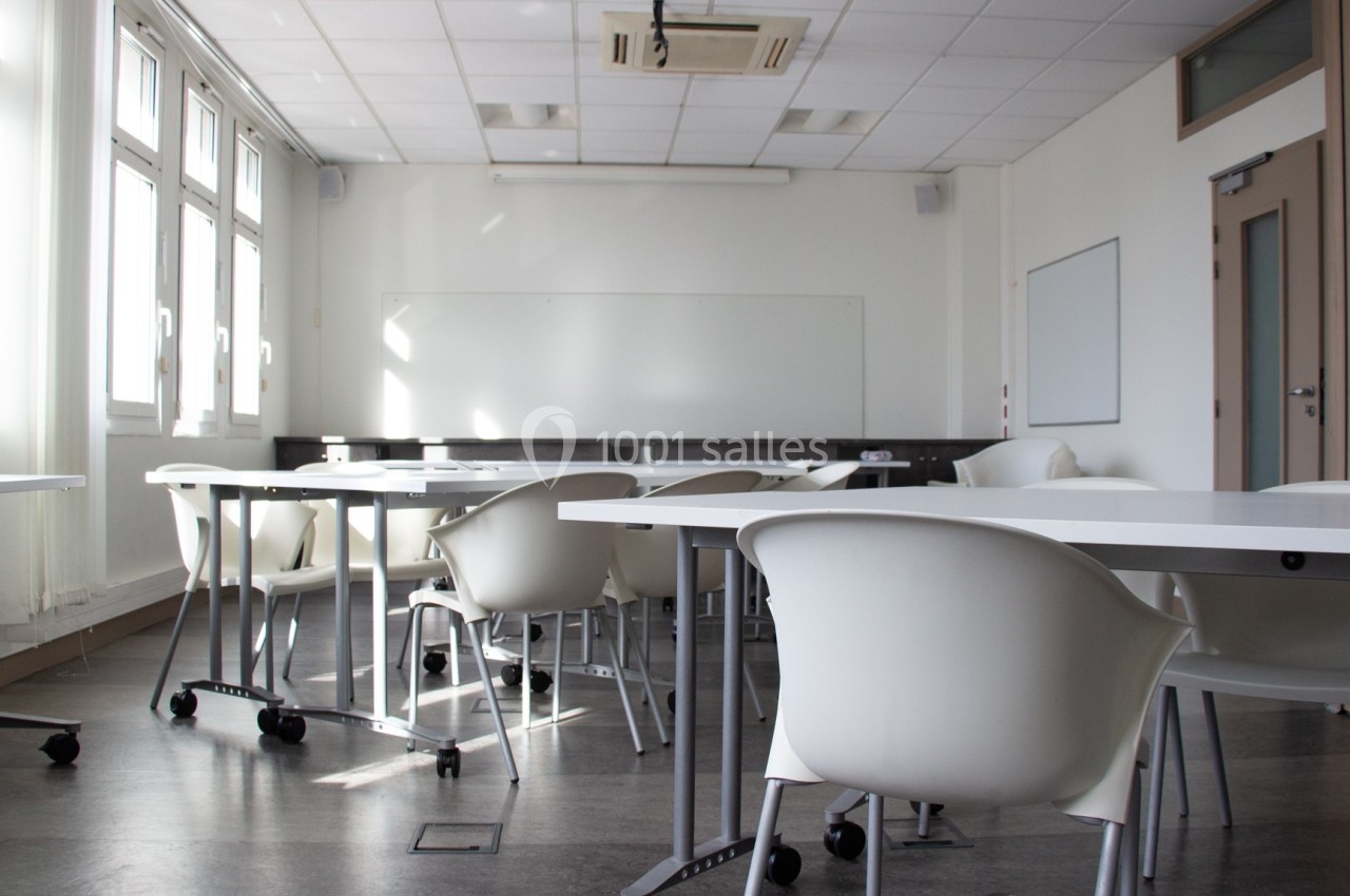 Salle de classe lumineuse avec des tables blanches, des chaises modernes et un tableau blanc au fond.