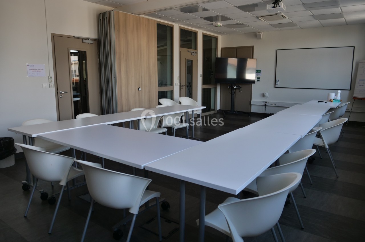 Salle de réunion vide avec des tables disposées en U, des chaises blanches et un tableau blanc au mur.