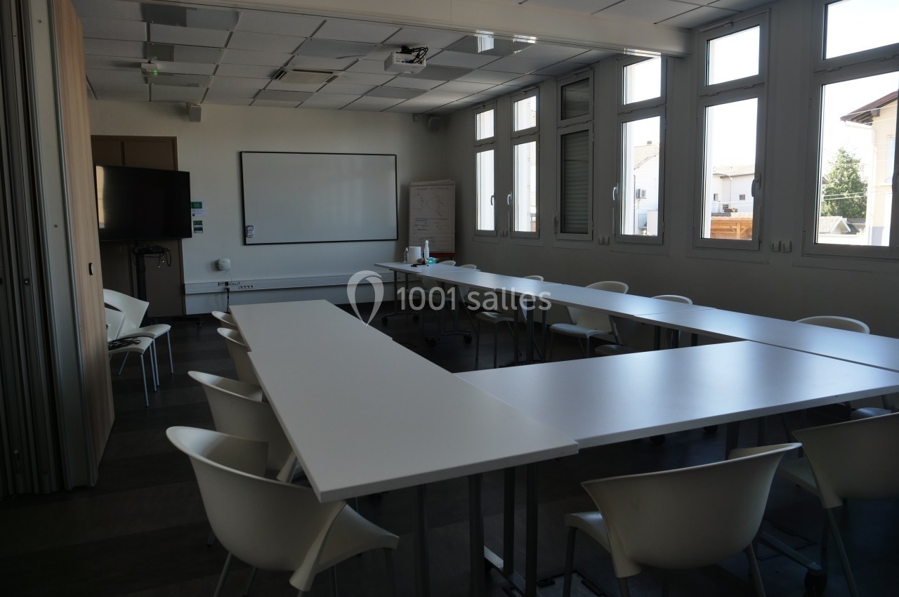 Salle de réunion lumineuse avec tables blanches en U, chaises, tableau blanc et écran mural.
